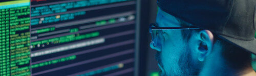 A person wearing glasses and a cap is working on a computer with lines of code displayed on the screen.