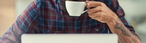 A person with a beard and tattooed arm wearing a plaid shirt drinks from a white mug while sitting at a laptop.