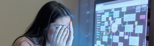 A person sits at a computer with hands covering their face, appearing stressed, while a complex flowchart is displayed on the monitor.