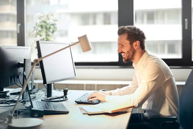 Man smiling while working at a computer in a bright office setting.