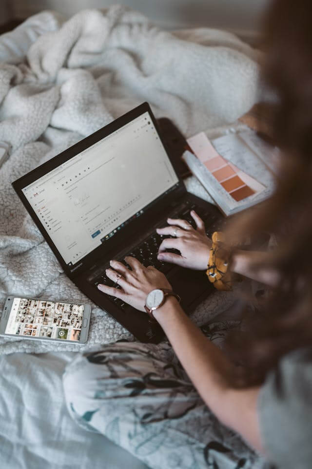 A person sitting on a bed and typing on a laptop with color swatches and a smartphone beside them.