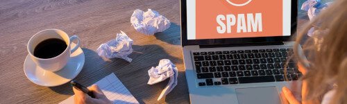 Person working at a desk with a laptop displaying "spam" message, crumpled papers, and a coffee cup.