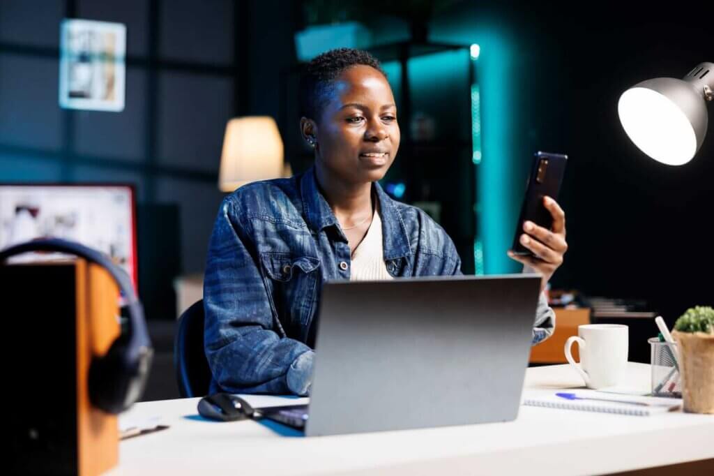 Person in a denim jacket sitting at a desk with a laptop, looking at their smartphone. A lamp, headphones, and a cup are on the desk.
