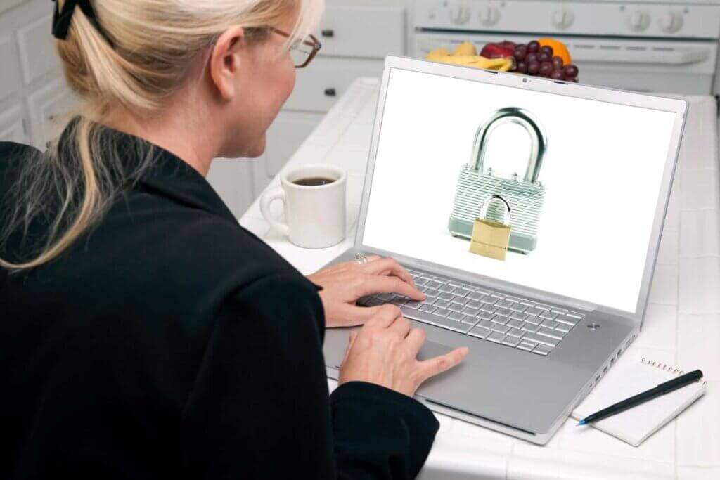 A woman in business attire uses a laptop displaying images of two padlocks, emphasizing email security. A cup of coffee, a pen, and a notepad are on the counter beside her.