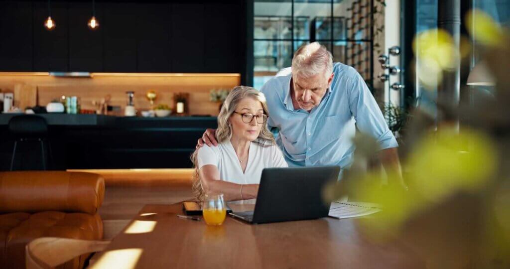 An older couple looks at a laptop together in a modern kitchen, with papers and a glass of orange juice on the table as they discuss email security.