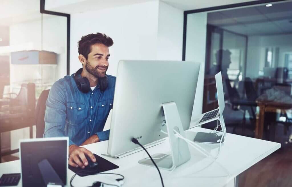 A man sits at a desk using a large computer monitor, with headphones around his neck and other electronic devices on the desk in a modern office setting, focused on email security tasks.