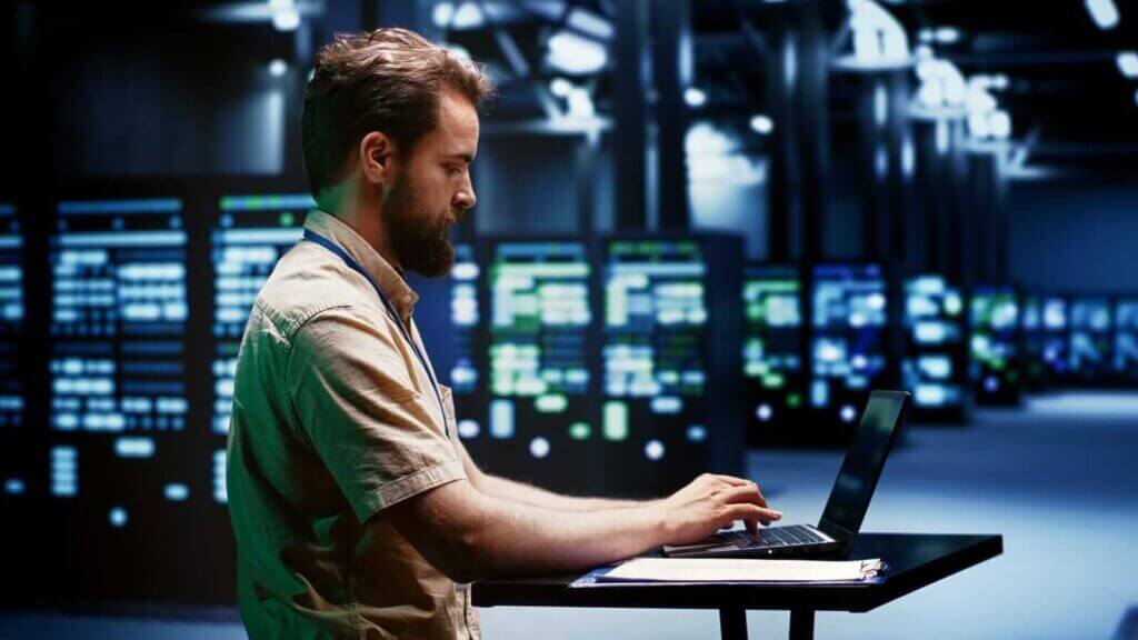 Man working on a laptop in a server room with multiple server racks and computer equipment, showcasing the robust infrastructure behind web hosting Singapore.