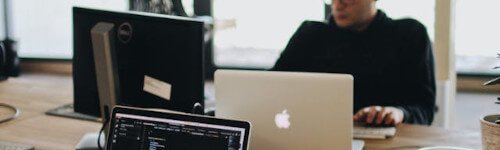 Two people work at a desk with laptops and monitors in an office setting, collaborating on web hosting Singapore solutions. One screen displays lines of code.