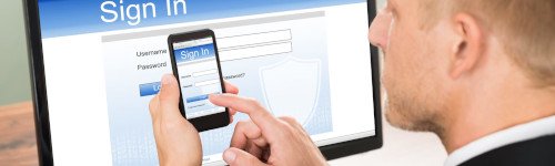A person uses a smartphone to sign in to a website displayed on a computer monitor, entering login credentials on both devices.